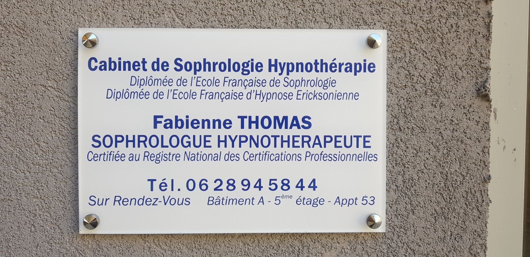 Panneau blanc d'un cabinet de sophrologie et hypnothérapie Fabienne Thomas sur un mur gris. Indique : Diplômée des Ecoles Française de Sophrologie et d'Hypnose Ericksonienne. SOPHROLOGUE HYPNOTHERAPEUTE. Tél. 06 28 94 58 44. Sur Rendez-Vous. Bâtiment A - 5ème étage - Appt 53.