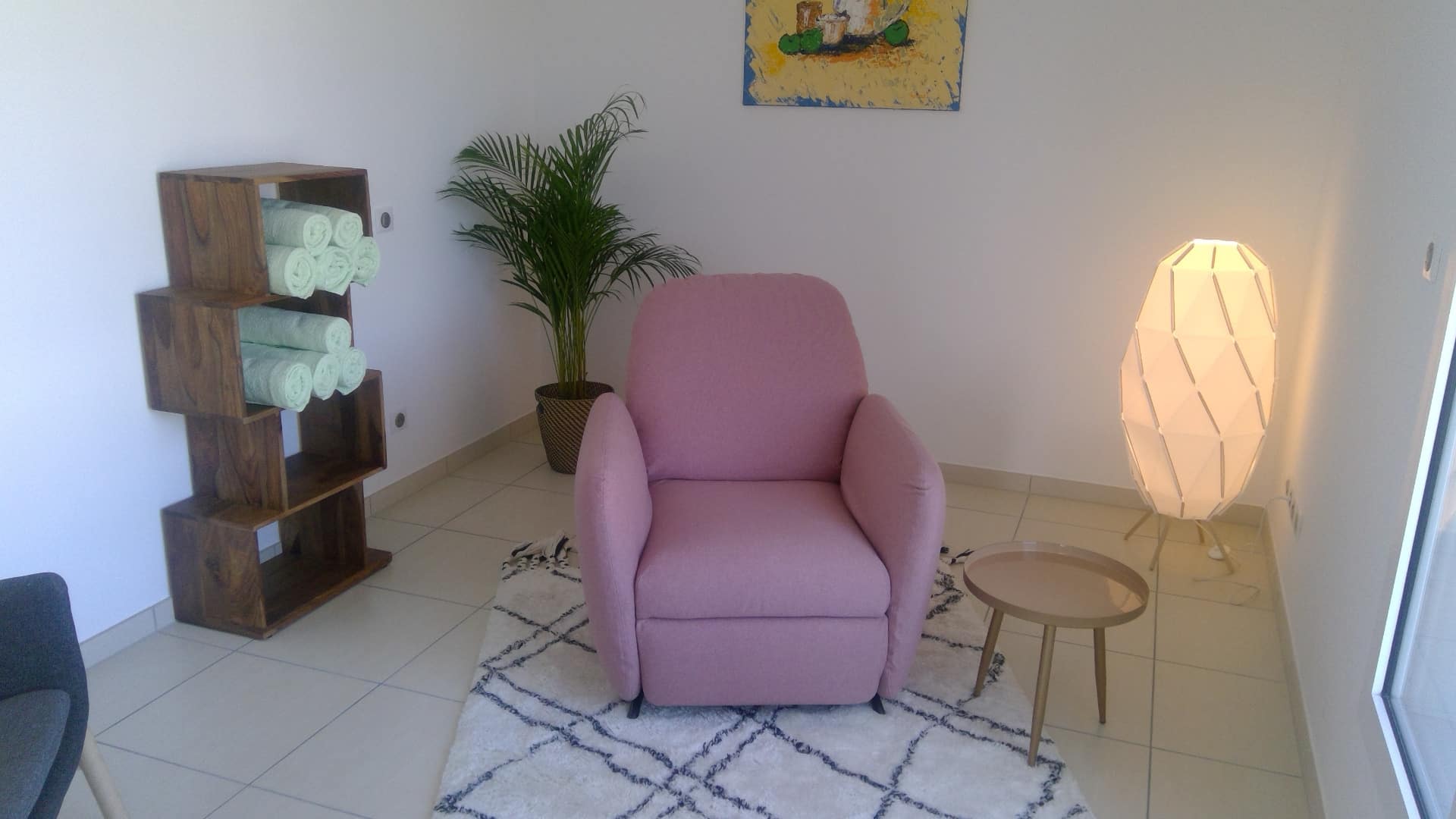 Fauteuil rose sur tapis géométrique, étagère bois avec serviettes, plante, tableau et lampe design. Espace accueil relaxant.