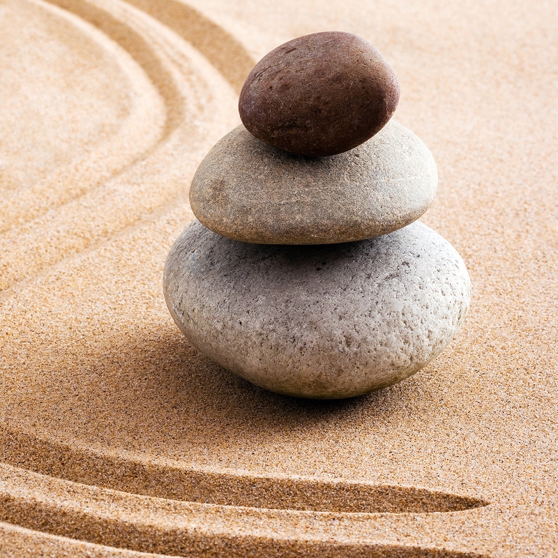 Galets Zen Empilés : Harmonie et Équilibre sur Sable Trois galets lisses empilés en équilibre sur du sable beige finement ratissé avec des motifs de jardin zen.
