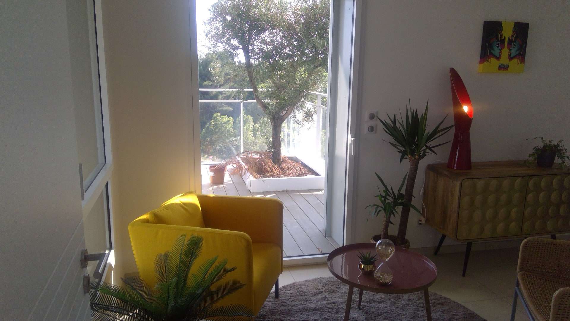 Salon moderne lumineux avec fauteuil jaune, meuble en bois et lampes. Accès à un balcon ensoleillé avec olivier et vue sur la nature verdoyante.