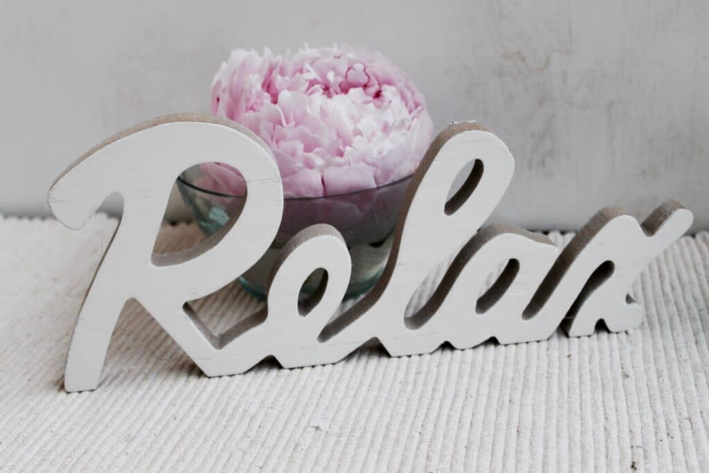 Détendez-vous : Signe 'Relax' et pivoine rose pour bien-être Signe en bois blanc 'relax' avec une pivoine rose dans un bol en verre sur une surface texturée blanche. Ambiance calme et reposante.